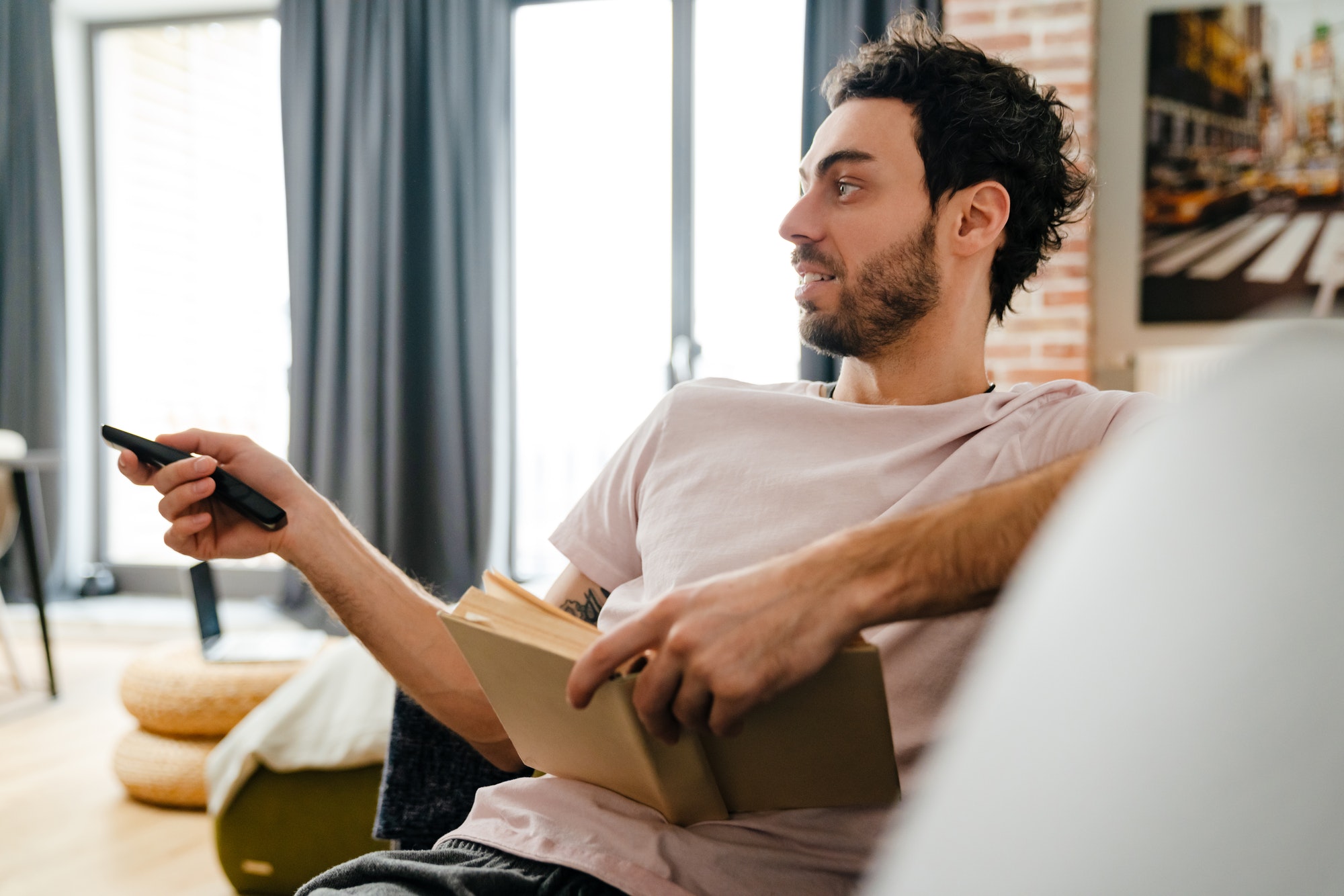 Handsome unshaven man using remote control while reading book on couch