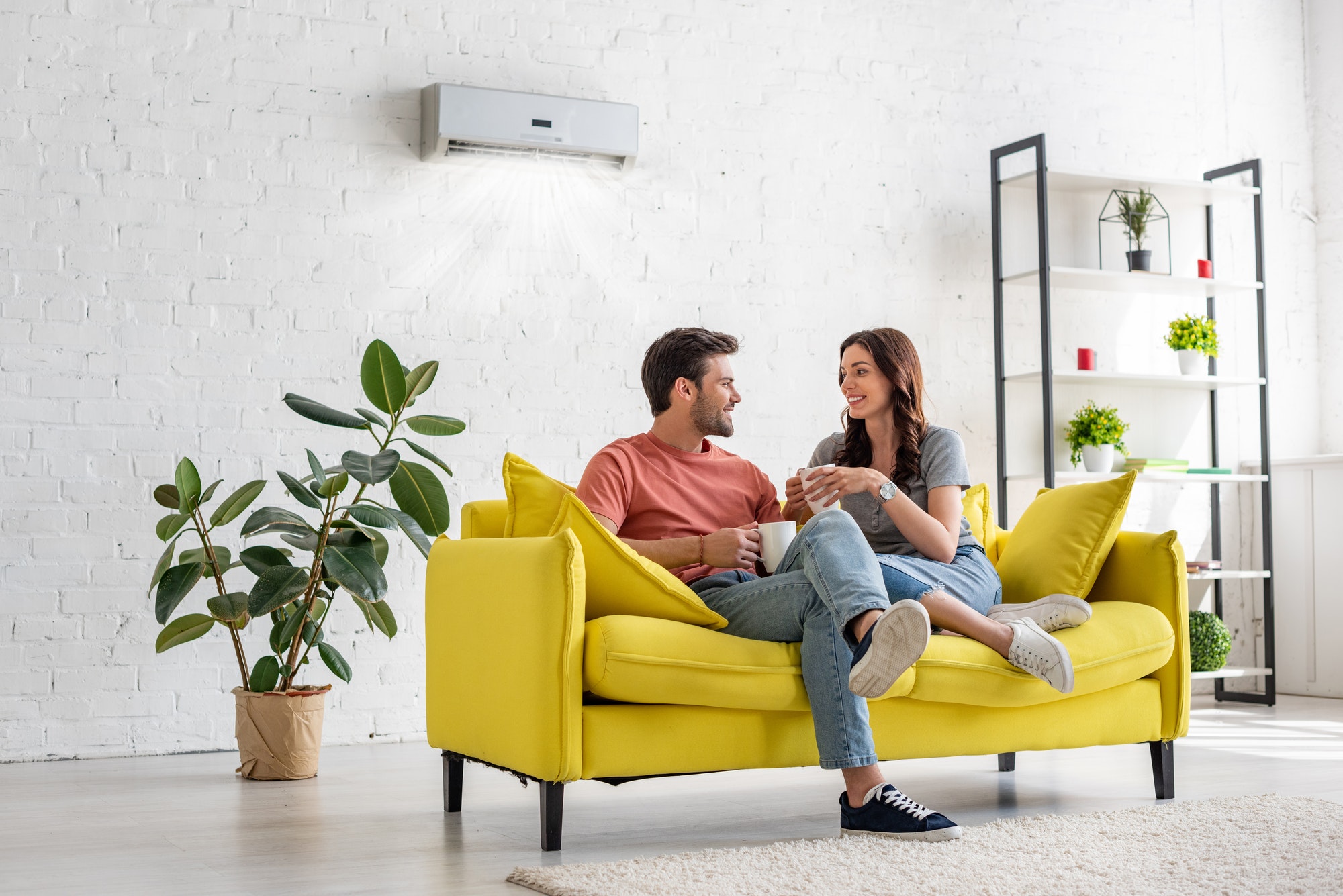 happy young couple talking and holding cups while sitting on yellow sofa under air conditioner at